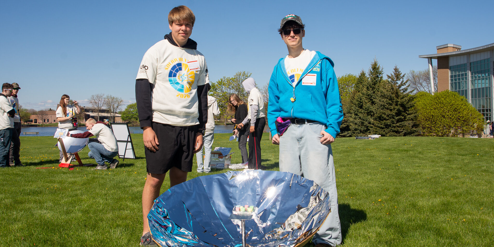 Two male students smile for a photo with a large reflective and circular dish harnessing solar energy at the 30th annual WPS Solar Olympics.
