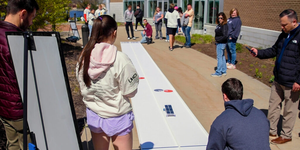 A small crowd of adults and high school students watches as a solar-powered model race car travels down a race track on a sunny day.