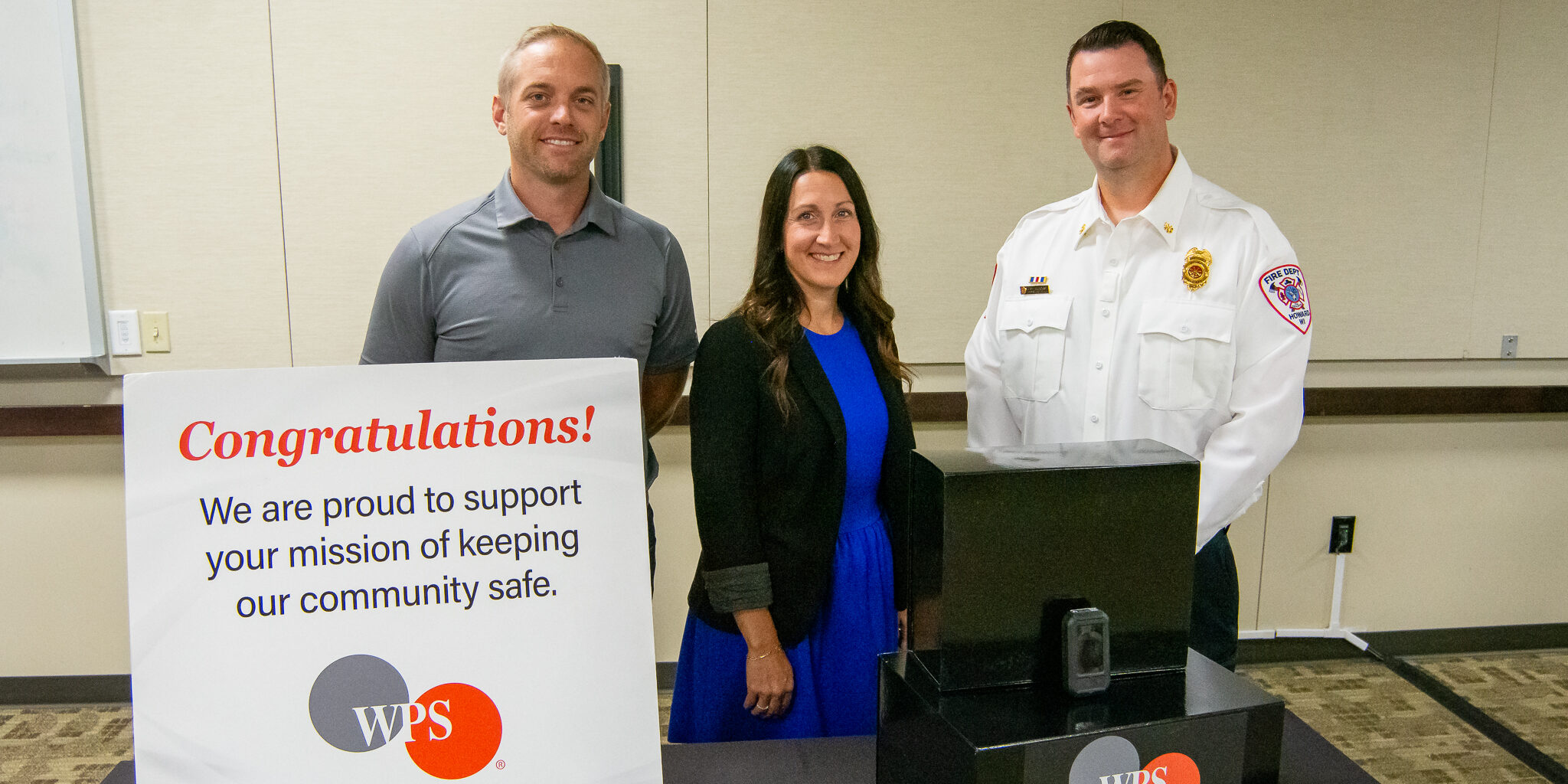 A man and woman from WPS join a Howard Fire Rescue firefighter for a photo behind a congratulatory sign and a small handheld radiation detection device.
