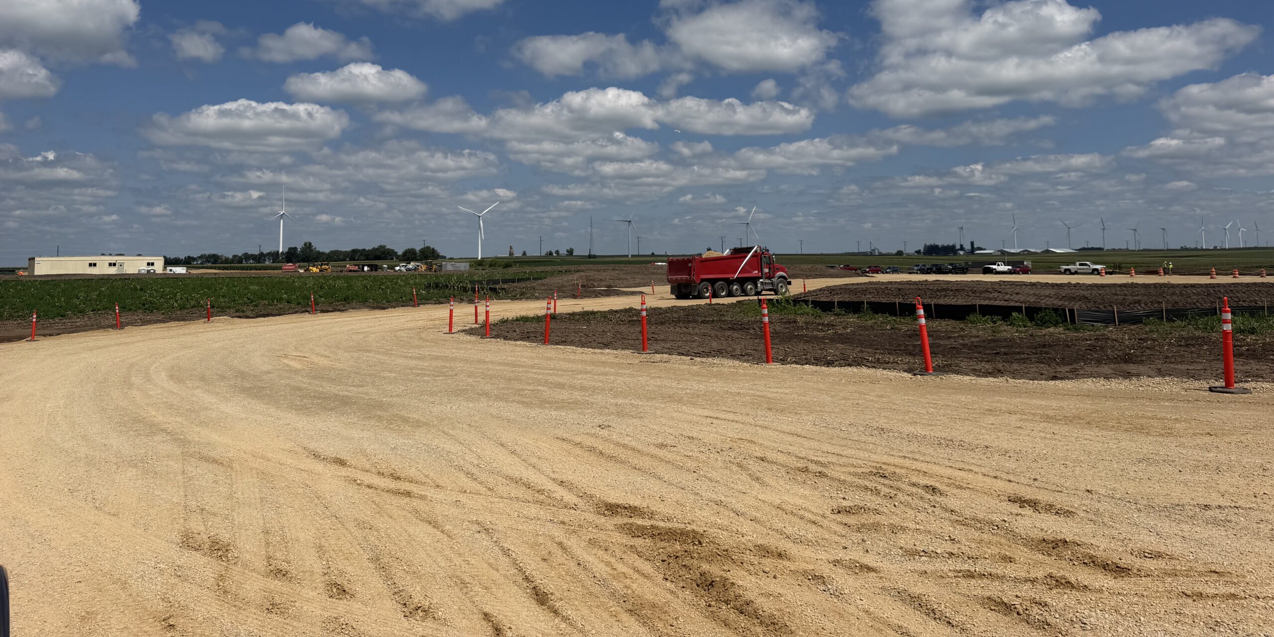 A photo of a truck driving on a dirt construction road with several wind turbines in the background.