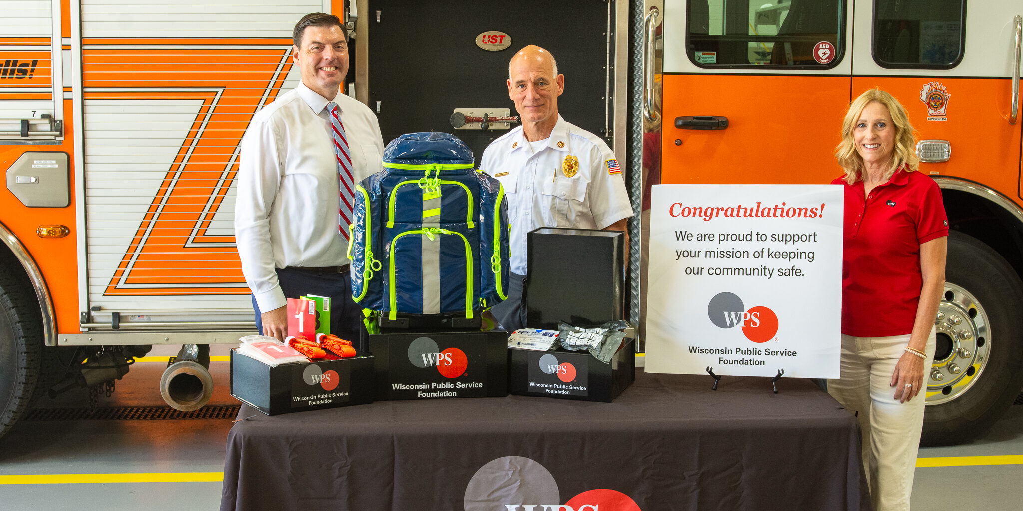 A man and woman with WPS stand on either side of a male fire chief behind a table with emergency medical supplies arranged on boxes with the WPS Foundation logo on them.