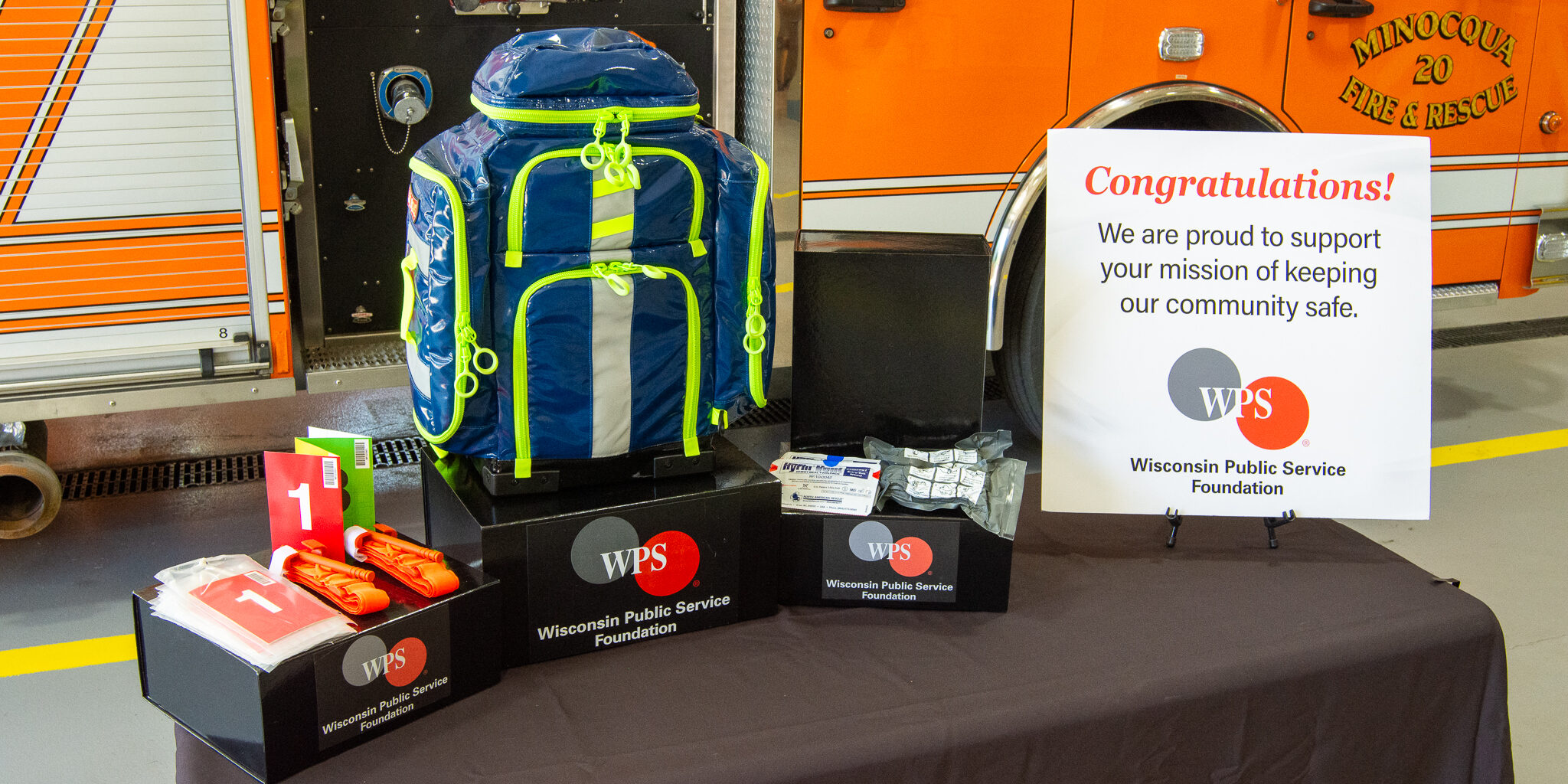 Emergency medical supplies and a blue and green backpack lying on top of boxes with the WPS Foundation logo on them. The boxes are next to a congratulatory sign from the WPS Foundation.
