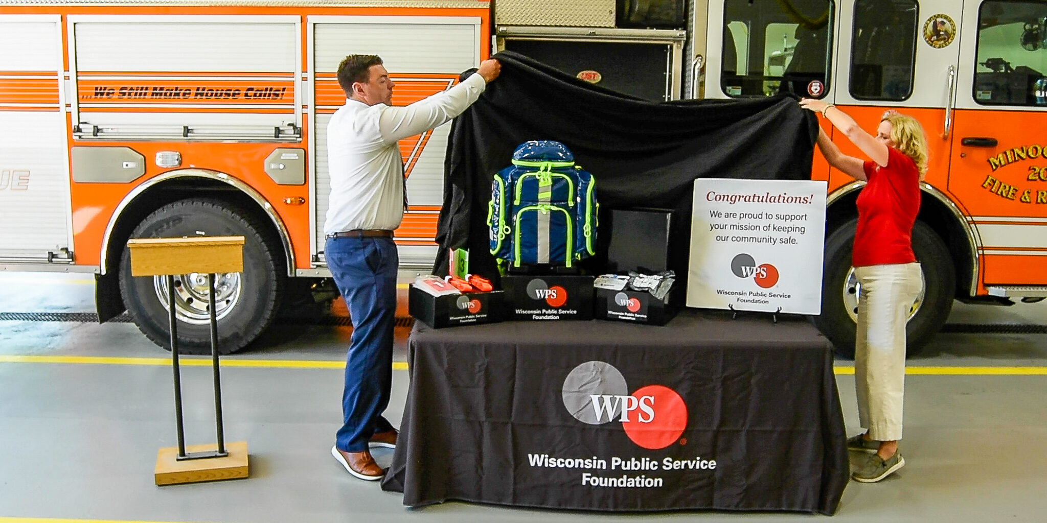 A man and woman with WPS help unveil emergency medical supplies arranged on a table covered by a WPS Foundation tablecloth.