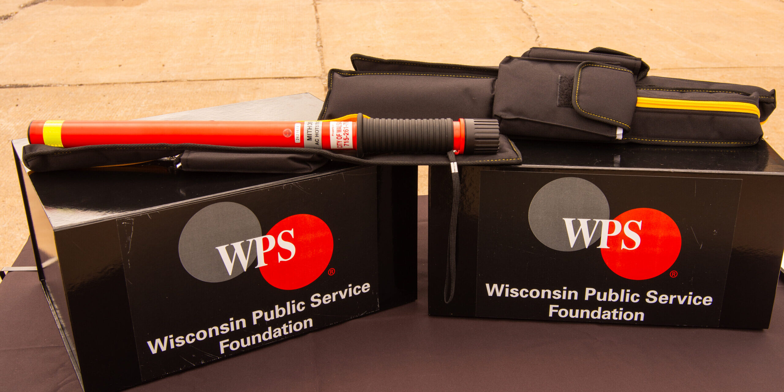 An orange electric hazard detection device lying near two more devices in protective cases. All three devices are lying on top of boxes with the WPS Foundation logo on the front.