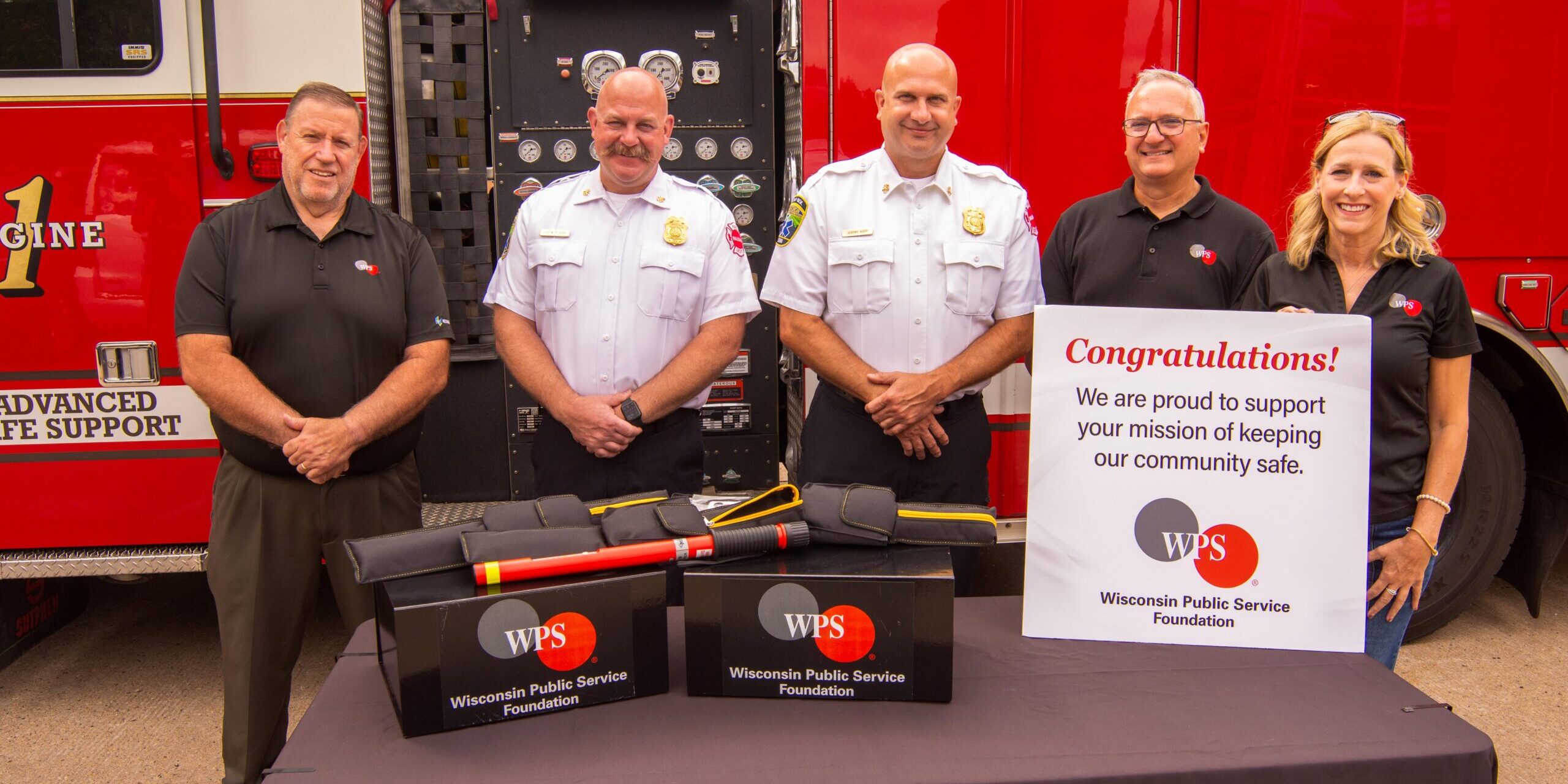 A photo of three WPS employees and two Wausau Fire Department firefighters standing in front of a fire engine. The group is standing behind a table with an electric hazard detection device lying on display boxes.