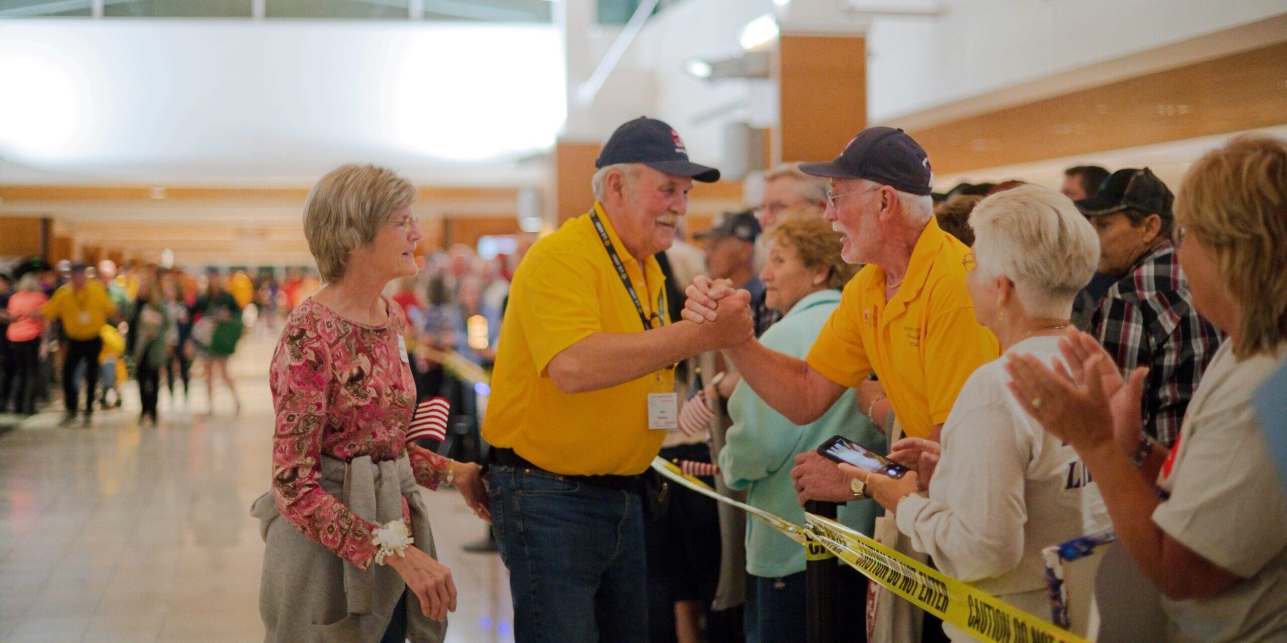 A military veteran wearing a yellow shirt is greeted by another veteran in a crowd of people at an airport.