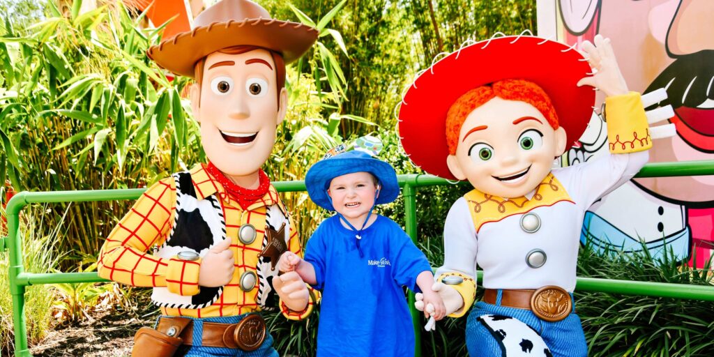 A young boy in a blue shirt smiles for a photo with the characters Woody and Jessie from the Toy Story movie franchise.