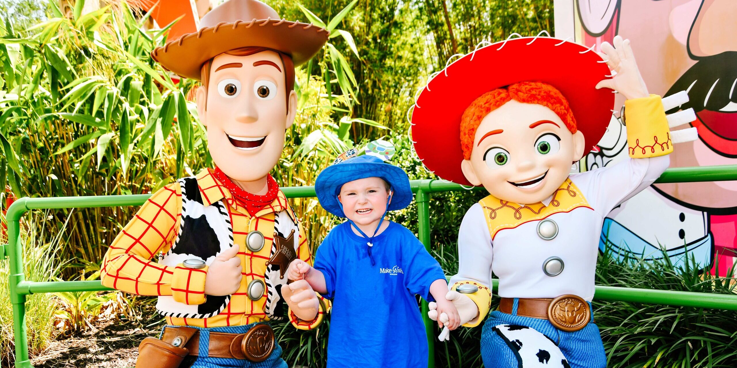 A young boy in a blue shirt smiles for a photo with the characters Woody and Jessie from the Toy Story movie franchise.