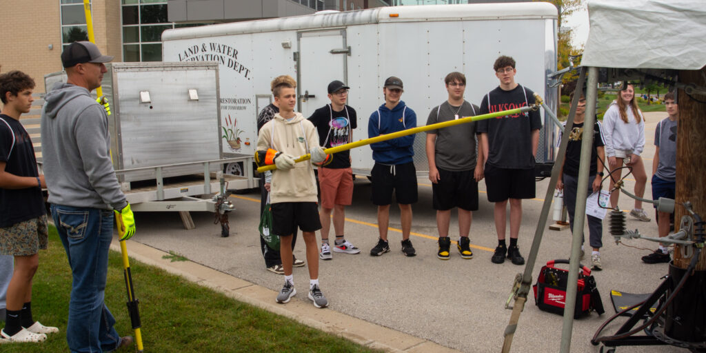 A high school student uses a long fiberglass stick to remove a mock fuse from an electric wiring demonstration in a parking lot.