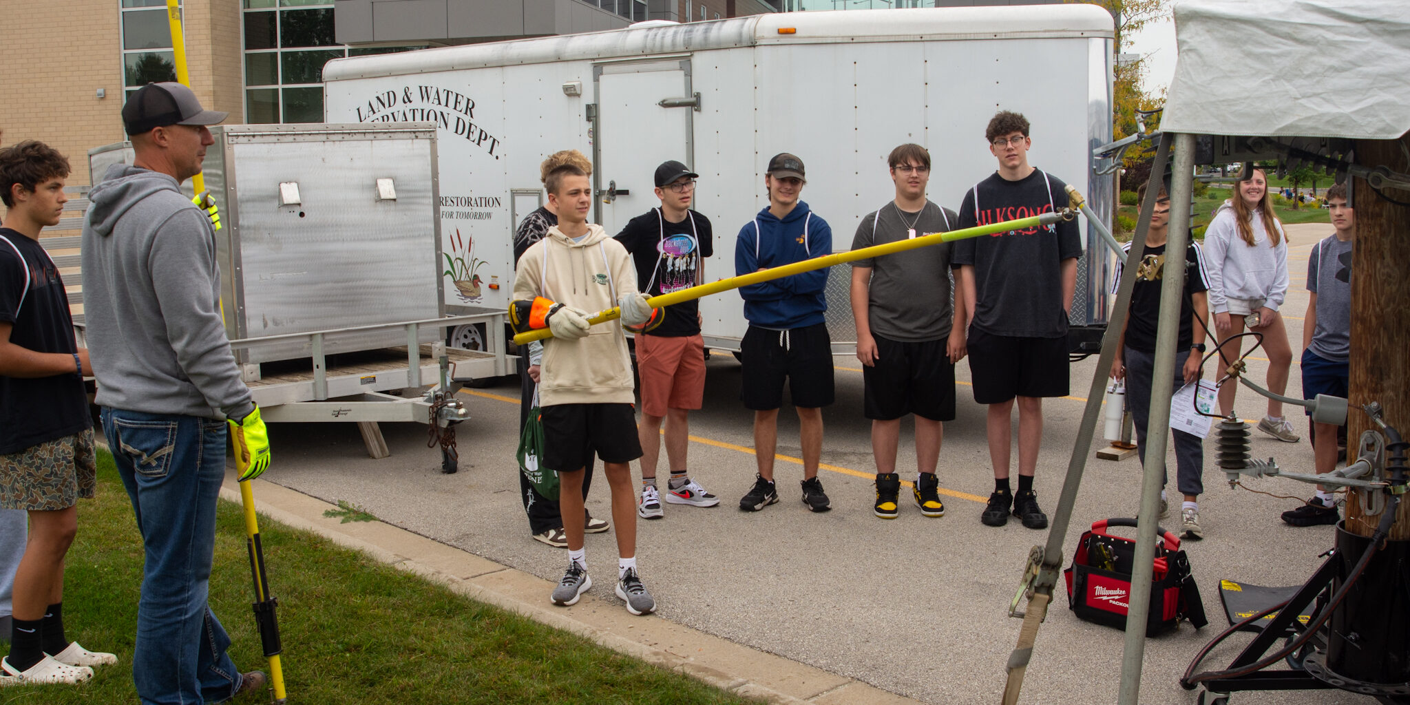 A high school student uses a long fiberglass stick to remove a mock fuse from an electric wiring demonstration in a parking lot.