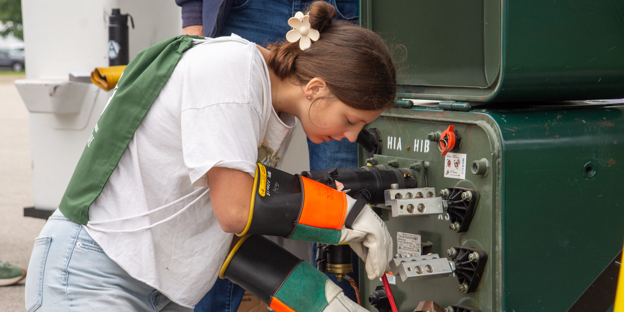 A female high school student works on a mock electric transformer while wearing heavy-duty rubber gloves.