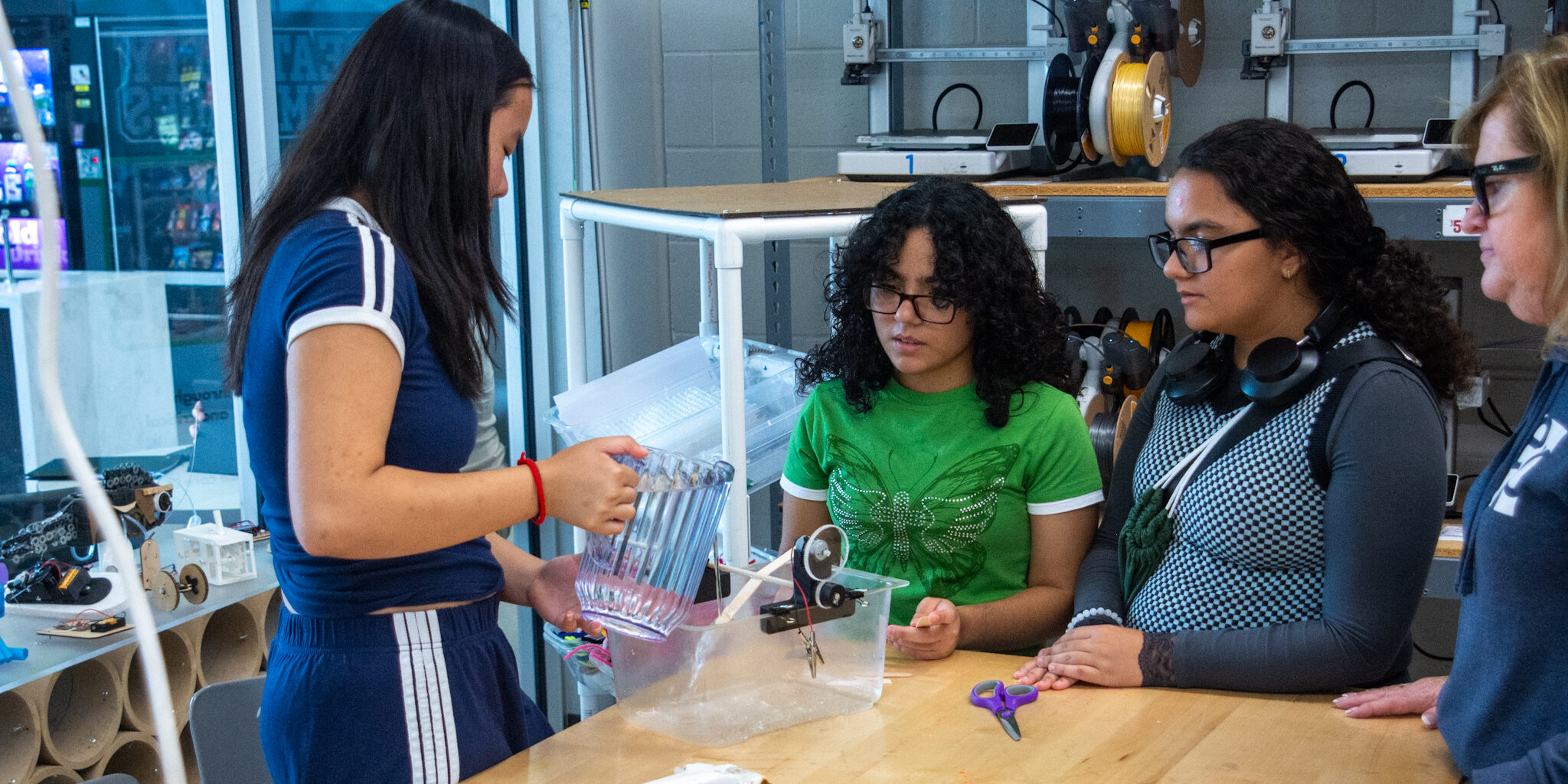 A small group of female high school students pours water from a pitcher onto a miniature hydroelectric power turbine.