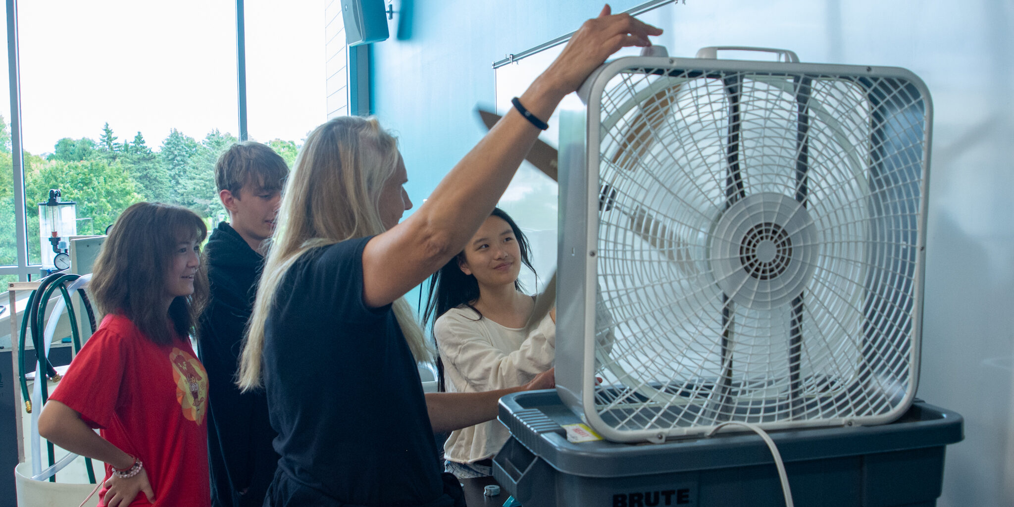 Three high school students test out a miniature wind turbine using a box fan with a an instructor inside a college lab classroom.